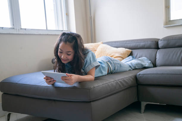 istock Young girl happily using digital tablet on a sofa at home 1474400247
