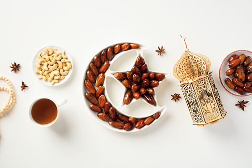 Ramadan Kareem concept. Islamic star and crescent bowl with dried dates, nuts, cup of tea, wooden oriental lantern, rosary on white background
