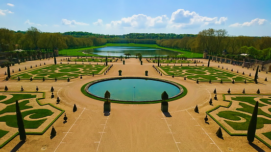 Estate of Versailles, Paris, France - April 20, 2018: aerial view of a designed garden in Versailles with trees, grass, a fountain and a lake in patterns under bright blue sky and clouds