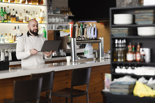 Side view of adult bearded male bartender in casual clothes smiling and looking at camera while working in modern bar