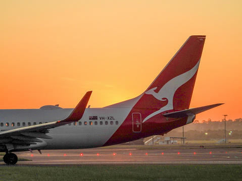 The vertical stabiliser of a Qantas Boeing B737-838, VH-XZL, flight number QF487, which is taxiing to the third runway of Sydney Kingsford-Smith Airport before taking off and heading to Melbourne.  This image was taken from Shep's Mound, a public viewing area off Ross Smith Avenue, at sunset on 18 March 2023.