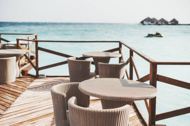 A tranquil outdoor cafe restaurant area filled with rattan-effect furniture, tables, and chairs in selective focus on the wooden promenade and fenced, in a peaceful background with a grey sky