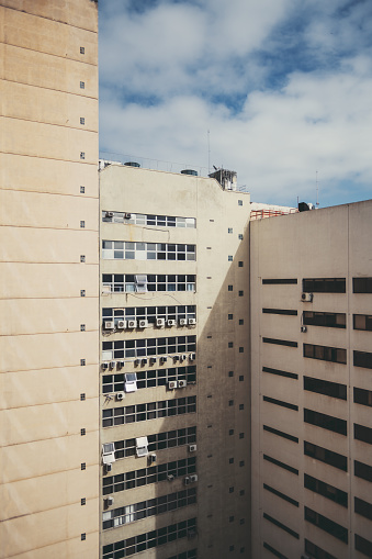A vertical capture of an inner corner of two yellow-to-cream-colored residential apartments with tiny squared windows, and many outdoor air conditioning unit compressors visible on a blue sky day