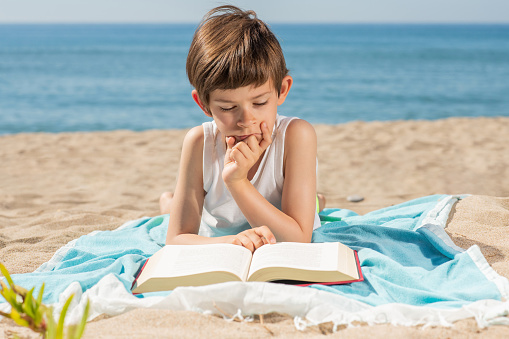 Child immersed in reading a book on the towel on the beach on a sunny day with the sea in the background