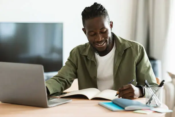 Smiling african american man sitting at desk, working on laptop and taking notes in notebook, black male studying online or watching webinar and writing check list
