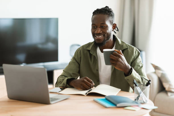 istock Happy black man drinking cup of tea at workplace while working on computer in home office interior 1474446976