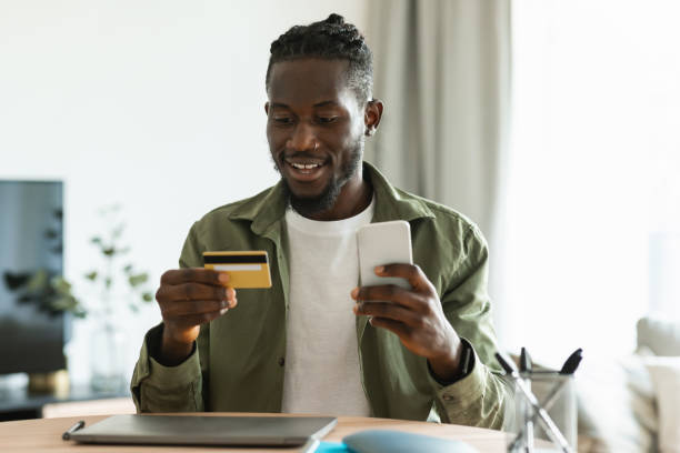 istock Internet mobile shopping. Happy black man using cellphone and credit card, purchasing online while sitting at desk 1474446985