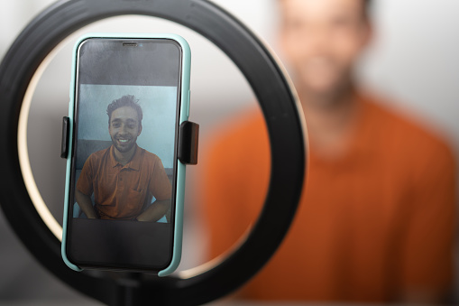 Smiling man wearing an orange polo shirt, taking a picture with his cell phone in a ring light.