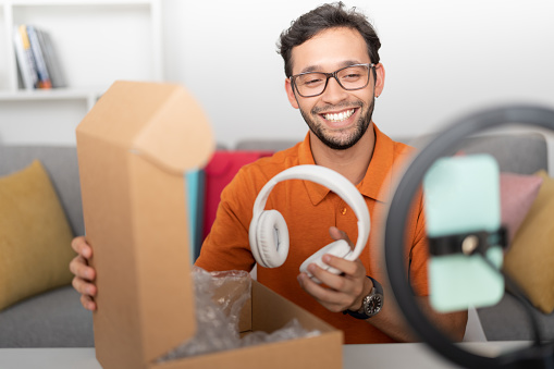 Male influencer wearing glasses and an orange polo shirt, sitting at a desk recording a video with his cell phone in a ring of light while unboxing a gift of white headphones.
