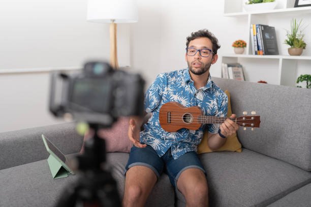 istock Man sitting in a living room, teaching ukulele playing while recording a video with a camera on a tripod. 1474468042