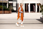 Street Fashion. Unique style for glamour woman walking city, wearing fashionable clothes white pants and loafers shoes, jacket, scarf and glasses. Side view