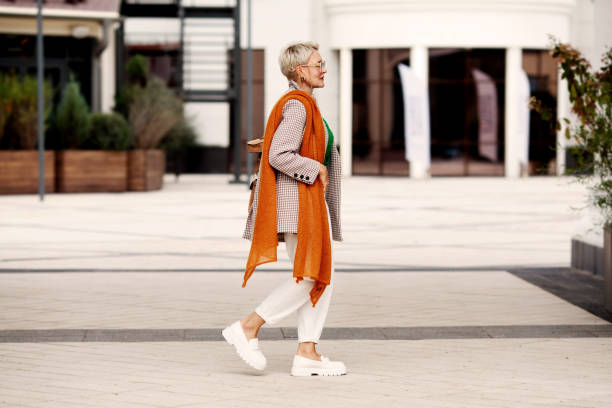 Street Fashion. Unique style for glamour woman walking city, wearing fashionable clothes white pants and loafers shoes, jacket, scarf and glasses. Side view stock photo