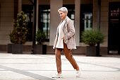 Full length stylish woman walking Street and wears trendy business clothes outfit, oversize jacket, tie, pants and white loafer shoes. Confident female model with short blonde haircut