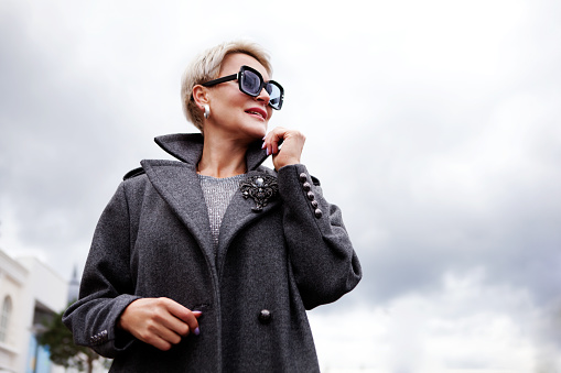 Portrait attractive female model on cloudy sky background. Modern stylish woman with short hair in gray double breasted pea coat and glasses