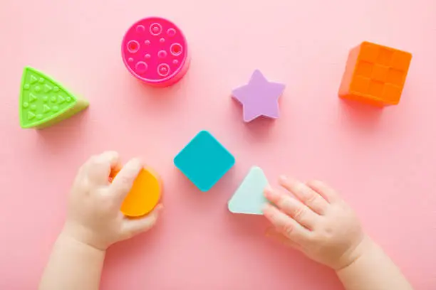 Photo of Baby girl hands playing with colorful plastic shapes on light pink table background. Pastel color. Closeup. Infant development toys. Point of view shot. Top down view.