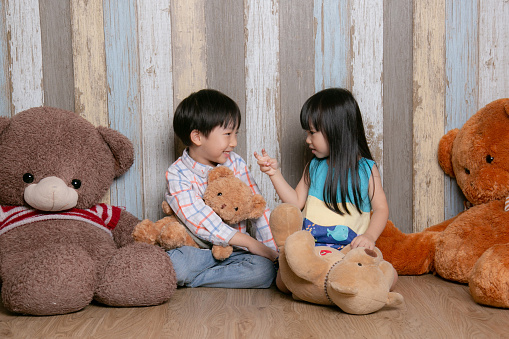 A boy and girl are engaged in a lively conversation while being surrounded by teddy bears. The boy is smiling while the girl gestures the number 2.