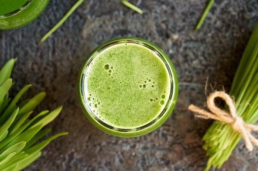 A glass of homemade green barley grass juice with freshly grown blades on dark background, top view