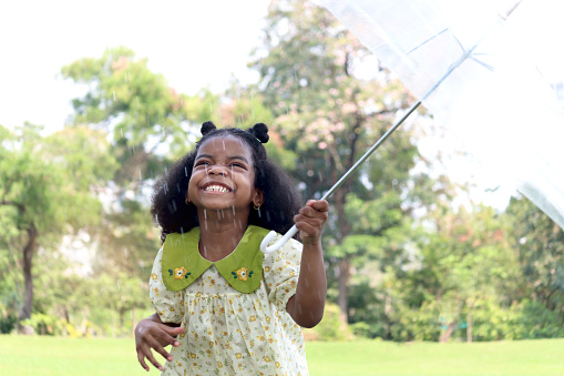 Happy smiley African girl with black curly hair holding umbrella under raindrops fall while standing outdoor green park, beautiful kid playing outside garden on rainy day, cute child playing in the rain.