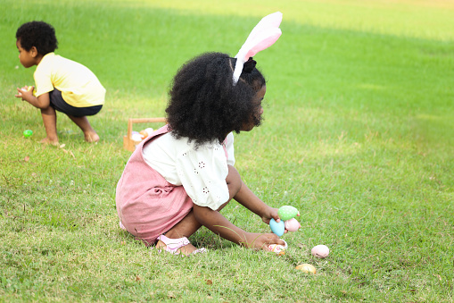 Happy African girl with black curly hair wearing bunny ears find, pick up Easter eggs hunt in green garden with her brother, kids celebrating Easter at outdoor park, friend children play outside