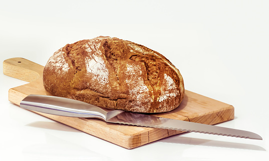 Studio shot of Rustic wholegrain brown bread loaf, on wooden cutting board with stainless steel bread knife, set on neutral white background.