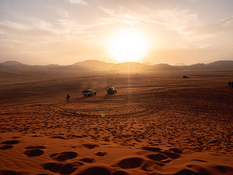 A view of the Wadi Rum desert with off-road vehicles in the sand, in the distance the sun is setting and people are approaching the parked vehicles.