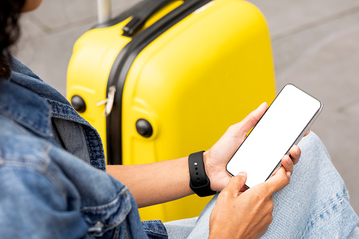Unrecognizable lady in casual outfit sitting on bench with yellow suitcase outdoors, using modern smartphone with white empty screen, using travel app, booking hotel, tickets, taxi, cropped, mockup