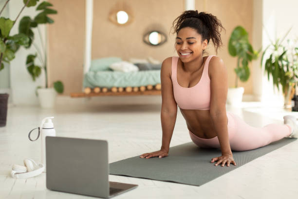istock Happy african american woman stretching back and watching online tutorials on laptop, excercising in bedroom on yoga mat 1474553771
