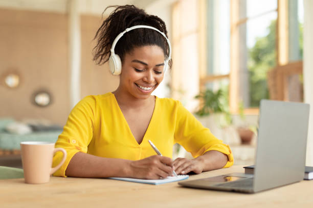 istock Happy african american lady studying remotely from home, using laptop and making notes, sitting at desk at home 1474554576