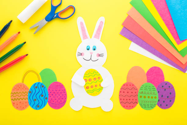 istock White smiling bunny and colorful eggs from paper with different supplies on bright yellow table background. Closeup. Making easter decoration elements. Top down view. 1474555340