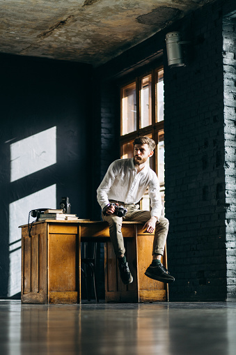 The handsome man sitting on the table and holding film camera in hist hands in creative studio