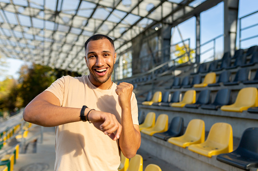 Young African American male athlete outdoors is happy with the result on the fitness bracelet. Shows a victory gesture with a yes hand, celebrates, smiles and looks at the camera.