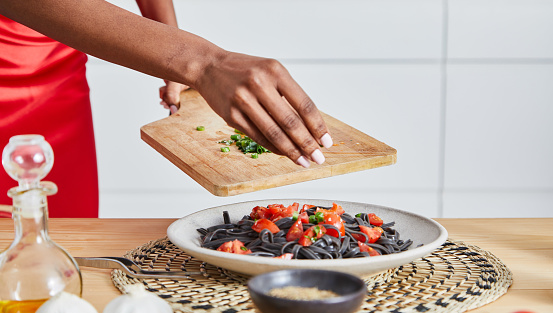 Beautiful young adult black woman preparing a healthy vegan pasta, made of black tagliatelle, cherry tomato, green onion and garlic, served on an elegant ceramic plate, with fresh vegetables and herbs, pumpkin seed and sesame seed, olive oil and pepper, on a light wooden natural kitchen or restaurant table, representing a wellbeing and a healthy lifestyle and food indulgence, image with a copy space