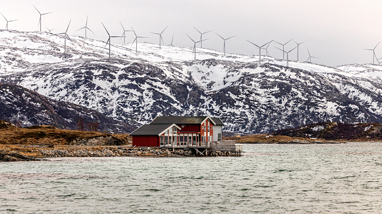 Red house by the sea with snowcapped mountains and high wind turbines on the top of snowcapped mountains.,Norway