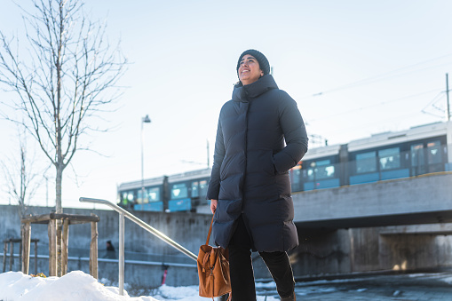 A female commuter in the city, she is at a railroad station on a sunny winter day. It's cold, the sun is shining and the ground is covered by snow.