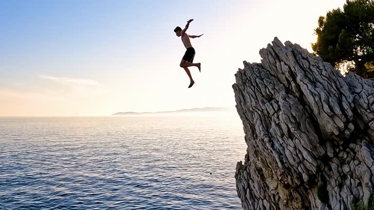 Young man jumping off cliff into the sea at sunset