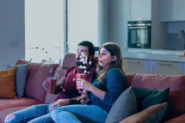 Photo of friends on couch watching TV - couple sitting on sofa scared the hell out of them watching horror movie - flying popcorn