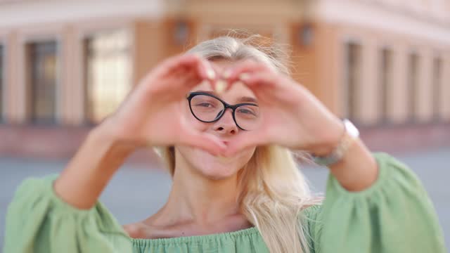 Close-up of sincerely smiling young blonde woman showing the camera a symbol of love- heart shape with fingers,  expressing romantic feelings, social work, donation charity concept