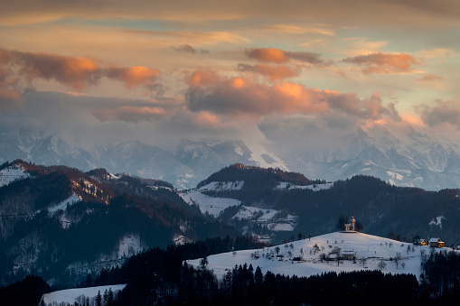Breathtaking Slovenian sunrise landscape with Julian Alps and charming little church of Sveti Tomaz (St. Thomas) on a hill in winter time, Slovenia