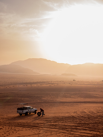 A beduin man changing a 4x4 tire in the middle of the Wadi Rum Desert while the sun is setting in the background.