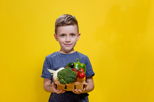 The boy is holding a basket with fresh vegetables on a yellow background. Basket with broccoli, cauliflower, cherry tomatoes, cucumber, pepper. Vegan and healthy concepts
