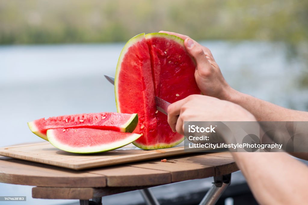 Cutting up Fresh Watermelon by the Lake Closeup of an anonymous person's hands holding and slicing up a watermelon fruit into pieces on a cutting board outside in the sun on a warm summer day. Watermelon Stock Photo