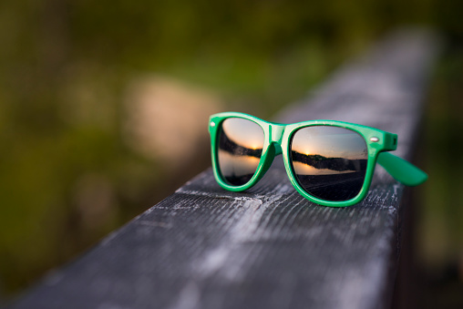 Macro closeup of vibrant, green coloured sunglasses sitting on a wooden deck with the reflection of the beautiful summer evening sunset and lake in the lenses.