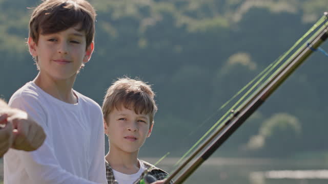 Fisherman teaches boys to cast fishing rod correctly. Fisherman teaches boys to cast rod correctly. People stand on river bank on summer day. Boys look at man with interest and older boy nods and smiles.