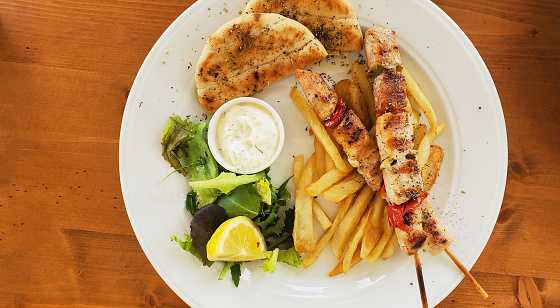 chicken skewers, french fries, sauce and bread tortilla on a white plate, garnished with lemon and sprigs of herbs. fast food in a cafe. delicious and satisfying food