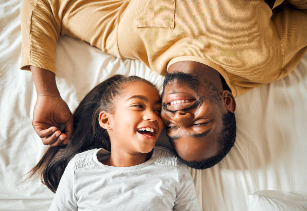 black family, father and daughter love on a bed with a smile on face or laughing at funny memory. above man and child together in home bedroom for bonding, support and care with safety and security - nostalgia joy laughing wellbeing imagens e fotografias de stock