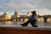 Stylish beautiful young woman wearing black hat sitting on Vltava river shore in Prague with Charles Bridge on background. Elegant retro lady fine art portrait.