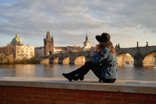 Stylish beautiful young woman wearing black hat sitting on Vltava river shore in Prague with Charles Bridge on background. Elegant retro lady fine art portrait. stock photo