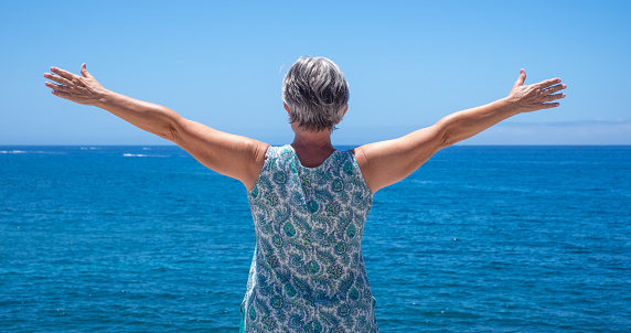 Rear view of caucasian elderly woman with outstretched arms in front to the sea looking at horizon. Freedom and summer vacation concept