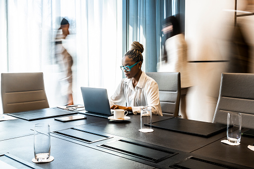 Busy hours in a board room during working hours while black contemporary businesswoman is looking at her laptop and her two colleagues are passing behind her in a conference room during business hours.