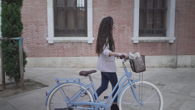 Curly-Haired Lady With Bicycle Is Walking Along The Sidestreet Of City Park. Tracking Shot